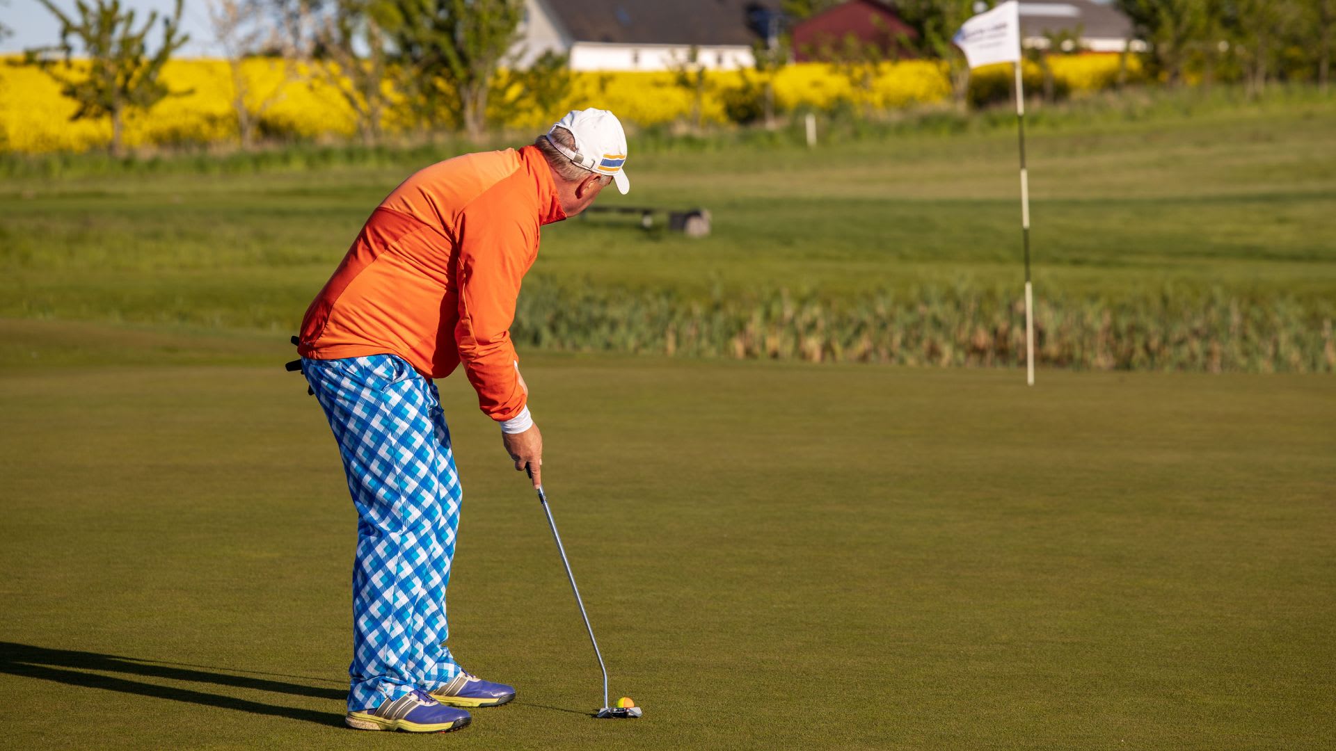 A golfer in a white cap, orange jacket, and blue patterned pants lines up a putt on a green with a flagstick in the background, perhaps pondering how long does golfing take on such perfectly manicured courses.